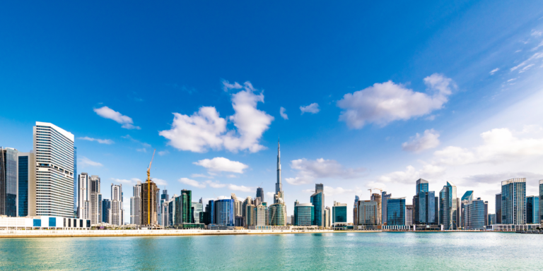 A panoramic daytime view of Dubai’s iconic skyline with the Burj Khalifa rising tall among modern skyscrapers, reflecting over clear blue waters under a bright sky — an essential highlight on any Dubai travel checklist, covering must-see landmarks, urban attractions, and unforgettable cityscape views.