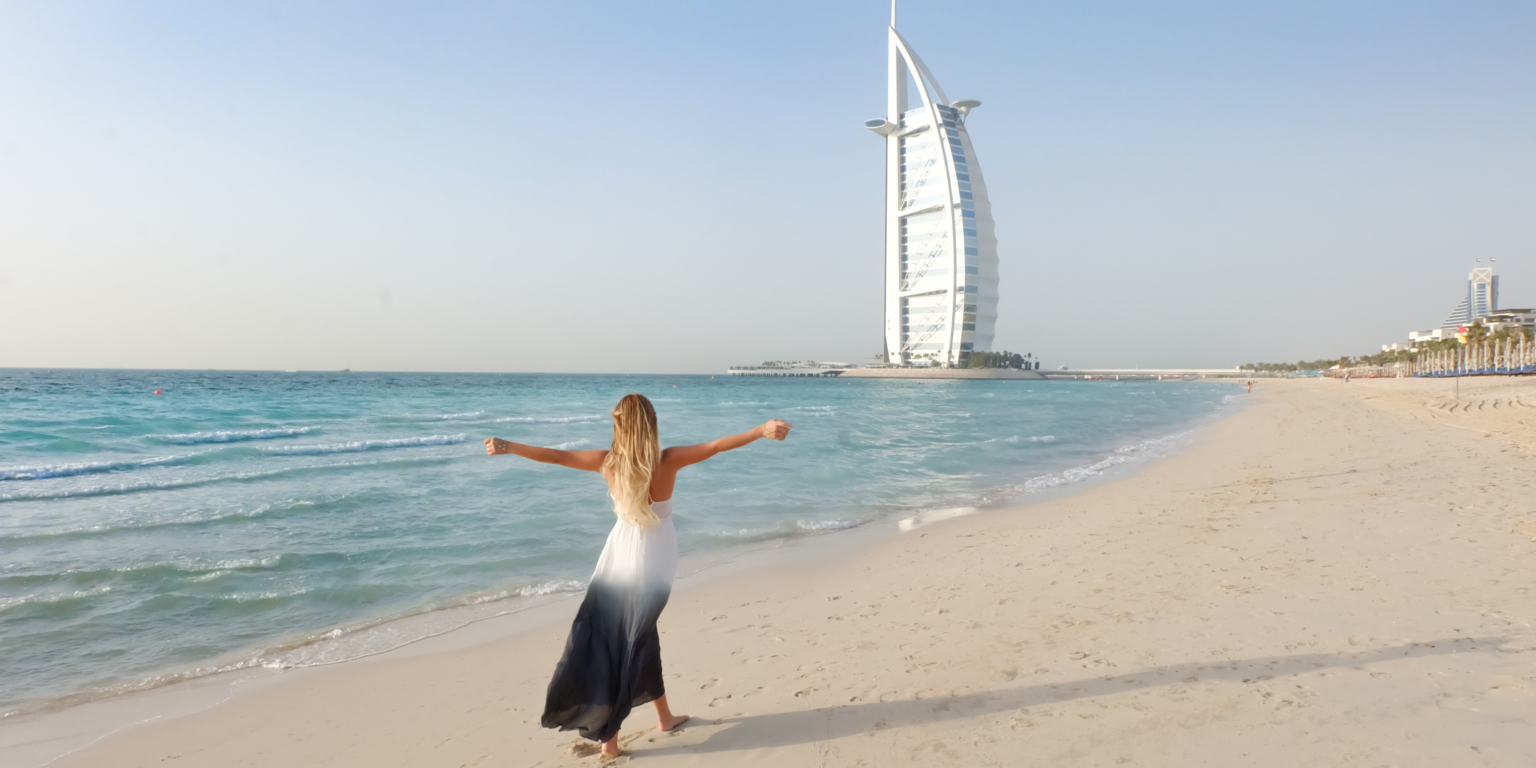 Woman in sleeveless dress walking on Dubai beach near Burj Al Arab showing how to dress in Dubai at the beach. the background shows burj al arab at a distance and the girl is enjoying the beach during the day time.